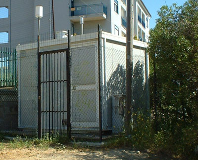 Cabina di rilevamento atmosferico di colore grigio sabbia ubicata in zona urbana. Alle spalle edifici di civile abitazione, in zona frontale area parco giochi.